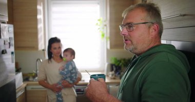 Baba bardağı tutarken anne arka planda bebeği taşıyor, aileyi gösteriyor ve mutfakta sakin bir ev havası var.