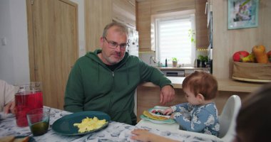 Babam, bebeğini renkli kahvaltı malzemeleriyle yüksek bir sandalyeye oturtup sıcak mutfak ortamında sıcak ve samimi bir aile ortamı yaratıyor.