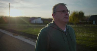 Gün batımında dışarıda yürüyen gözlüklü orta yaşlı bir adam, rahat ve düşünceli bir yüz ifadesi, kırsal alan, akşam ışığı, sakin atmosfer, kırsal huzur.
