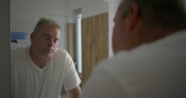 Aynada yansımaya bakan bir adam, rutin tımar sonrası düşünceli bir yüz ifadesi, beyaz tişört ve doğal banyo düzeni, sakin ve yansıtıcı atmosfer.