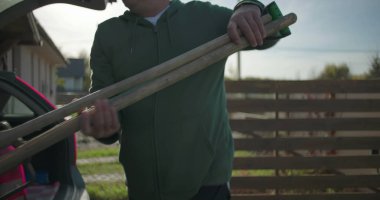 Bir adam arabanın bagajından kürek çıkartıyor, bahçe ya da açık hava görevlerine hazırlanıyor, güneşli bir konut bahçesinde çaba ve pratikliği vurguluyor.