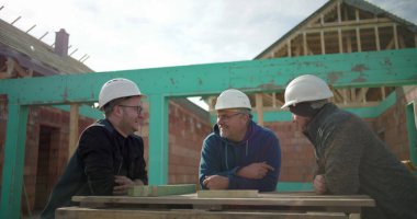 İnşaat işçileri, arka planda tamamlanmamış bir yapı iskeleti olan bir inşaat sahasında proje planlarını tartışırken tasasız bir anı paylaşıyorlar.
