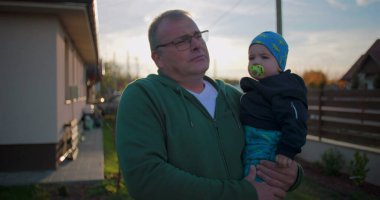 Father holding baby with pacifier in backyard, gently adjusting child's outfit, showcasing nurturing bond and love in a serene family environment