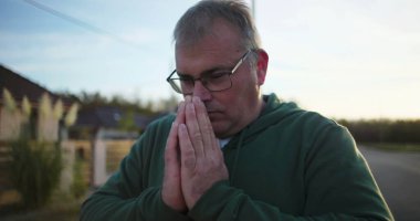 Dışarıda dua eden, elleri sımsıkı bağlı, aşağıya bakan, derin düşüncelere dalmış, günbatımında sakin kırsal bir sahne, minnettarlık ve maneviyat, huzurlu bir atmosfer.