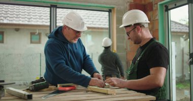 Çalışma tezgahındaki odunları ölçen ve hazırlayan inşaat işçileri, takım çalışması ve hassasiyet, güvenlik kaskları, marangozluk ve inşaat işleri, kapalı inşaat alanları,