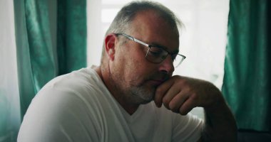 Çenesini elinin üzerine koyan, düşünceli bir ifadeyle aşağıya bakan, kapalı bir ortamda pencerenin yanında oturan bir adamın yakın çekimi.