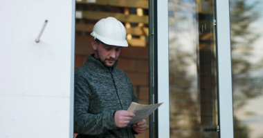 İnşaat işçisi kapıdaki planlar üzerinde çalışıyor, başlık takıyor, tamamlanmamış inşaat projesinin detaylarına odaklanıyor, inşaattaki hassasiyet ve kararlılığı aktarıyor.