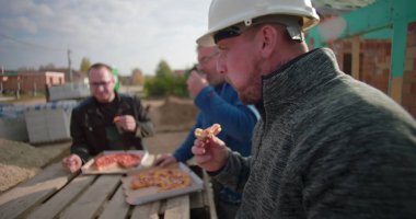 İnşaatçılar şantiyede pizzayla öğle yemeği arası veriyorlar, iş günlerinde gündelik anlar, takım çalışması ve dostluk, açık hava yemeği, konut projesi devam ediyor.