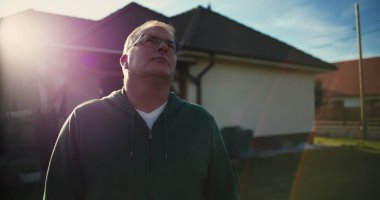 Evin dışına bakan bir adam, gözlük takıyor, banliyö bahçesinde huzurlu bir an, sakin bir ifade, yansıtıcı tavır, güneşli bir gün.