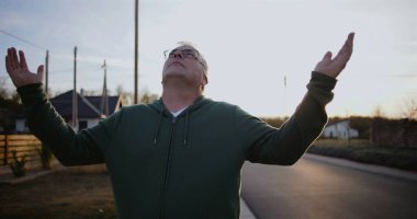 Dışarıdaki adam kollarını havaya kaldırmış, yukarıya bakıyor, minnettarlığını ve umudunu ifade ediyor, kırsal alanda ruhani bir an, sakin ve sakin, günbatımı atmosferi.