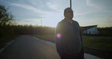 Orta yaşlı bir adam gün batımında kırsal bir yolda yürüyor, elleri ceplerinde, yansıtıcı ifade, huzurlu kırsal manzara, elektrik hatları, sakin alacakaranlık, kırsal ortam