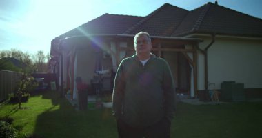 Arka bahçesinde gururlu bir ev sahibi olarak duran bir adam. Banliyö evi ve bakımlı bahçesiyle. Mülkünden ve yaşam tarzından gurur duyuyor.