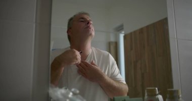 Boynunun altını tıraş ederken aynaya bakan, traş bıçağı ile kusursuz darbeler, doğal banyo geçmişi, aile ortamında kişisel bakım rutini