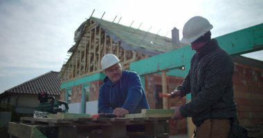 İnşaat işçisi ahşap çekiçlerken, meslektaşı inşaat alanında gözlem yapıyor, konut inşaatında takım çalışması ve el işçiliği yapıyor, arka planda ev iskeleti yapıyor.