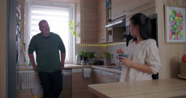 Mutfak tezgahına dayanmış bir adam elinde kahve fincanı tutan bir kadınla konuşuyor. İyi aydınlatılmış, ahşap iç mekânlı, modern bir mutfakta sıradan bir sohbet.