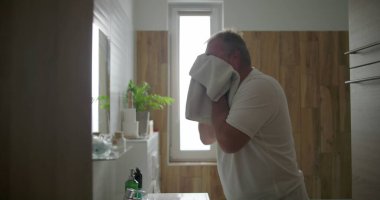 Beyaz havluyla yüzünü kurutan adam, duruladıktan sonra banyo lavabosunda duran, sabah bakım rutini, doğal ışık ve arka planda görünen ahşap banyo detayları.
