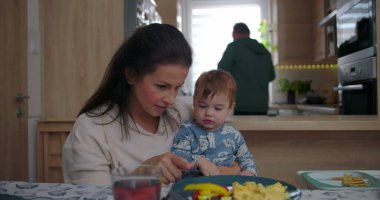 Anne yemek masasında bebeği besler, evde sıcak ve samimi bir aile ortamında renkli yiyeceklerle ilgilenir..