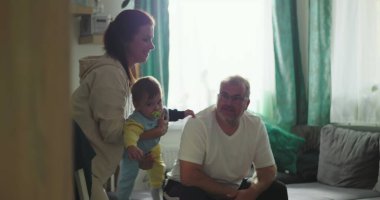 Anne kucağında bebek, baba rahat bir oturma odasında koltuktan izliyor. Neşeli aile bağları, bebek bakımı ve sıcak bir ortam.