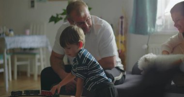 Baba ve oğul sehpanın üzerinde oyuncak trenle etkileşime giriyor, oyun saatleri ve bağlar paylaşılıyor, anne arka planda gülümsüyor, sıcak ev ortamında sıcak aile bağlantısı