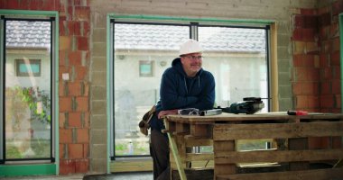 İnşaat işçisi kısmen inşa edilmiş bir yapıda, kollarını kavuşturmuş, kaskını takmış, inşaat alanındaki görevine odaklanmış.