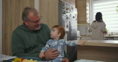 Baba mutfak tezgahında kucağında bebek, arka planda anne yemek hazırlıyor, sıcak aile anı, sabah rutini, birliktelik ve aşk bir arada..