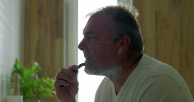 Banyoda tıraş olan bir adam, çene hizasında jilet kayması, tıraş köpüğü sürmesi, yan profil görünür, ayna yansıması, sabah bakım ritüelinin bir parçası.