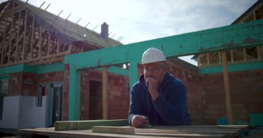 İnşaat işçisi inşaat alanında proje düşünüyor, eli çenesinde, ev yapısı ve arka planda çerçevesi olan düşünceli bir erkek inşaat işçisi.