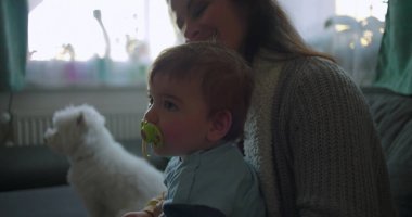 Toddler with pacifier sitting on mother's lap, looking curiously to side, fluffy white dog in background, relaxed family moment indoors