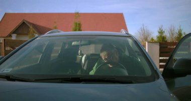 Bir adam konut yolunda sürücü tarafından arabaya biniyor, sürmeye hazırlanıyor, bir yolculuğa ya da her gün işe gidip gelmeye odaklanıyor