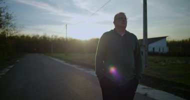 Orta yaşlı bir adam gün batımında kırsal bir yolda yürüyor, elleri ceplerinde, yansıtıcı ifade, huzurlu kırsal manzara, elektrik hatları, sakin alacakaranlık, kırsal ortam