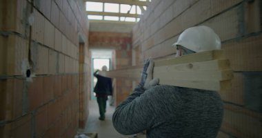 İnşaat işçisi inşaat halindeki konut binasında geniş ahşap kalasları taşırken, işçilere odaklanma ve inşaat ortamında çaba sarfediyor.