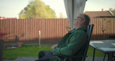 Avlu sandalyesinde oturan bir adam gözleri kapalı, sakin ve rahat görünüyor, çevresindeki yeşilliklerle huzurlu bahçe manzarasının tadını çıkarıyor.