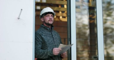 İnşaat işçisi kapıdaki planlar üzerinde çalışıyor, başlık takıyor, tamamlanmamış inşaat projesinin detaylarına odaklanıyor, inşaattaki hassasiyet ve kararlılığı aktarıyor.