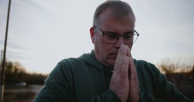 Elleri dua, yan görüş, yansıtıcı ve barışçıl ifade, sakin kırsal ortam, gün batımında ruhani an, şükran ve umut içinde kapalı bir adam.