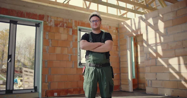 Kendine güveni tam bir inşaat işçisi, kolları çapraz, sıkı bir şapka ve tulum giyiyor, bitmemiş bir binanın içinde duruyor, kararlılık ve profesyonellik sergiliyor.