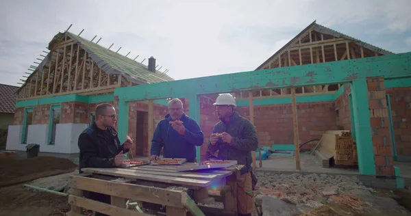 İnşaat ekibi şantiyede pizza molası veriyor, arka planda bitmemiş bir bina, günlük açık hava yemeği, inşaat projesi sırasında dinlenme anı, işçiler arasında dostluk.