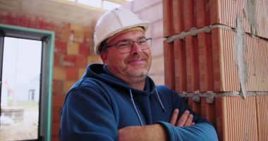 İnşaat işçisi inşaat halindeki tuğladan duvara yaslanıyor, kollar çapraz, güvenlik kaskı takıyor, rahat ve kendinden emin duruş, endüstriyel ortam.