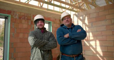 İki inşaat işçisi, kısmen inşa edilmiş bir binada güven içinde duruyor, kasketli şapkalar takıyor ve gülümsüyor, şantiyede takım çalışması ve güven gösterisi yapıyorlar.