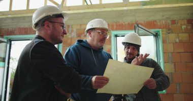 İnşaat ekibi tamamlanmamış binanın planlarını inceliyor, proje detayları, güvenlik kaskları, sanayi ortamında takım çalışması ve planlama üzerine odaklanmış tartışmalar yapıyor