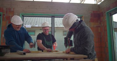 Ahşap işlerinde işbirliği yapan üç inşaat işçisi, ahşap masa üzerinde matkap ve aletler, inşaat alanında takım çalışması, güvenlik kaskları, kapalı alanda yenileme