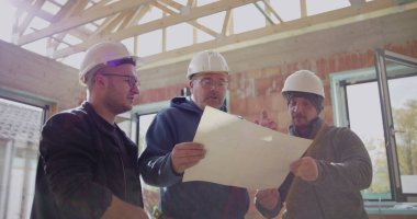 İyi aydınlatılmış bir inşaat alanının planlarını inceleyen inşaat işçileri, mimari planlara işaret ederek, proje detayları ve planlama üzerinde işbirliği yaparak,