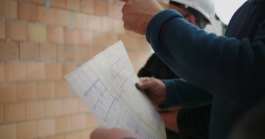 İnşaat planlarını, miğferleri, takım çalışmasını ve işbirliğini tartışan inşaat işçilerinin ayrıntılı görüşü, plan üzerinde yoğunlaşmak, inşaat alanı