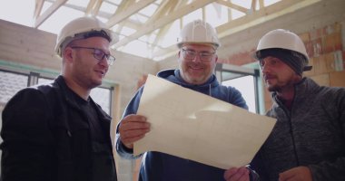 Üç inşaat işçisi gülümseyip bina planlarını tartışırken, inşaat projesiyle ilgili kısmen inşa edilmiş bir yapının planlarını gözden geçiriyorlar.