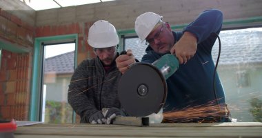 Metal üzerinde elektrikli öğütücü kullanan inşaat işçileri kıvılcımlar üretiyor, iş başında takım çalışması yapıyor, güvenlik kaskı ve koruma için gözlük takıyor, yenileme sahasını yeniliyor.