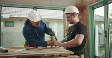 Tahta üzerinde elektrikli matkap kullanan iki inşaat işçisi, takım çalışması ve inşaat alanı görevi, güvenlik miğferleri, çevre inşasında işbirliği