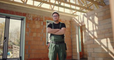 İnşaat işçisi güvenle duruyor. Kolları bağlı, kaskı ve tulumuyla inşaat alanında duruyor. İnşaat alanında gururlu ve profesyonel duruş sergiliyor.