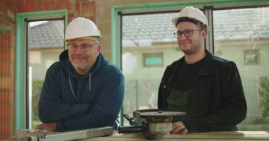 Çalışma masasında yan yana duran iki inşaat işçisi, güvenlik kaskı takıyorlar, takım çalışması ve inşaat alanında işbirliği yapıyorlar.