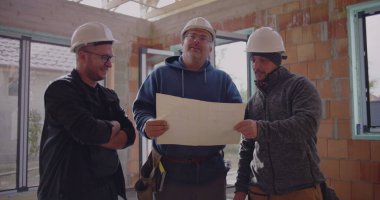 Üç inşaat işçisi planlarını gözden geçiriyorlar, güvenlik kaskı takıyorlar, proje detaylarını tartışıyorlar, takım çalışması ve bitmemiş endüstriyel alanda işbirliği yapıyorlar.