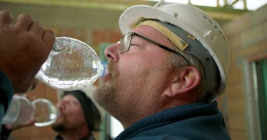 Mola sırasında su içen inşaat işçisi, susuz kalan, güvenlik kasklı adam bitmemiş bir şantiyede şişe suyla yenileniyor, profil görüntüsü yakın.