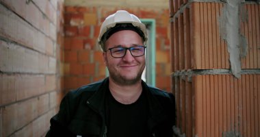 Kask ve gözlük takan, inşaat halindeki tuğladan duvara yaslanan, rahat ve dost canlısı tavırlar sergileyen gülümseyen inşaat işçilerinin yakın çekimi.